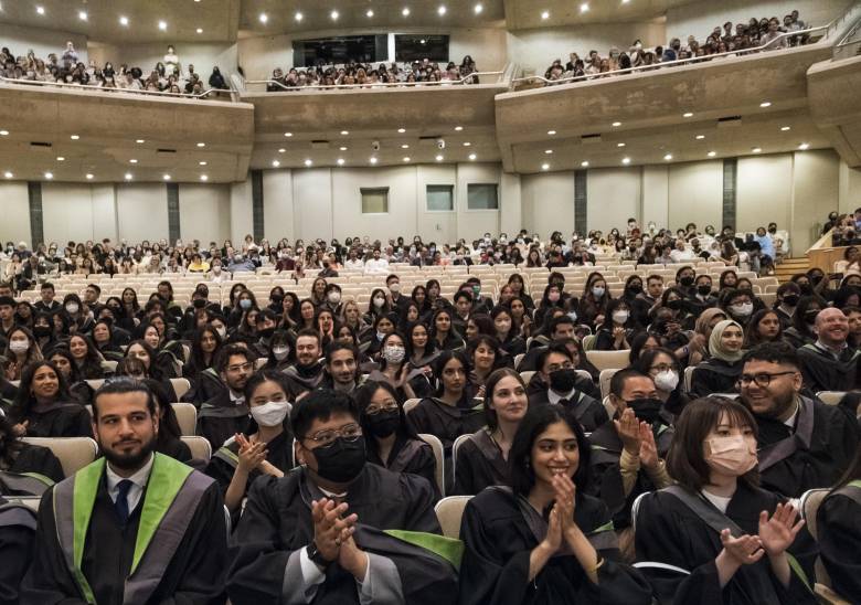 A group of students sit in an auditorium wearing black gowns and Convocation regalia.