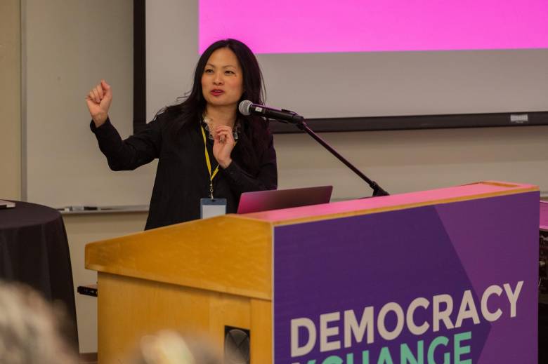 Associate Professor Ranee Lee speaks at a large podium with a microphone. The front of the podium has a sign with a purple background and white writing that reads DEMOCRACY XCHANGE.
