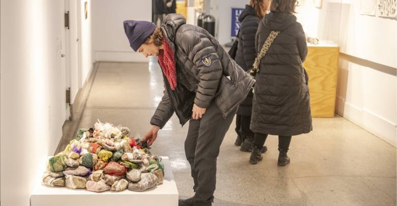 A person in a black jacket, navy toque and red scarf bends to touch an artwork with wrapped items on a stand. Two other people stand in the background looking at other artworks in the hallway.