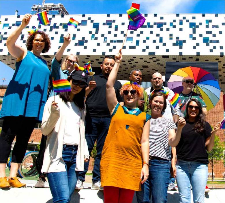 A group of people stand outside 100 McCaul St. on a sunny day, waving Pride flags and with other Pride-themed items, like an umbrella and unicorn ears.