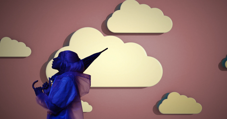 A person cast in a blue light stands in front of a backdrop in red with illustrated clouds.