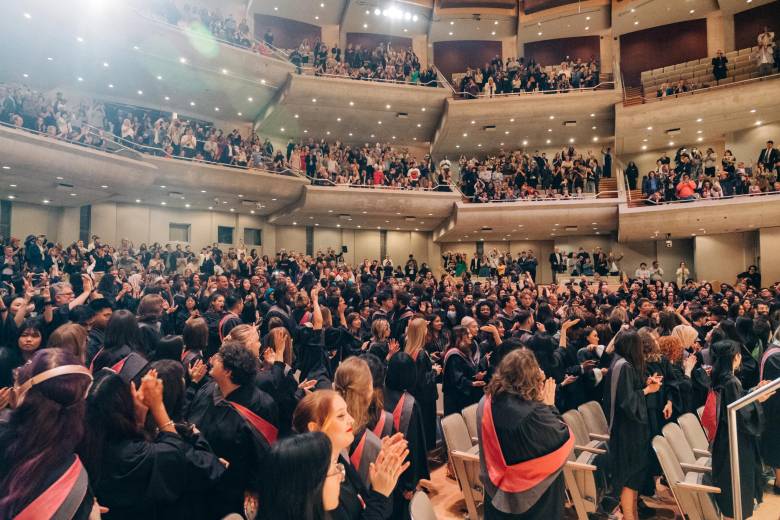 Lively photo from 2023 Convocation shows graduands in their robes, family members and friends in the audience, standing, clapping, smiling.