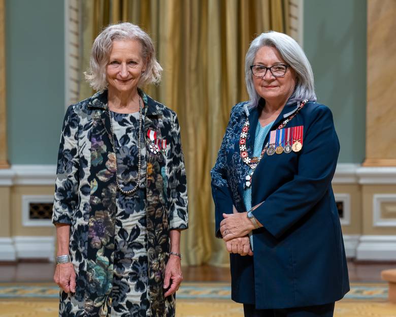 Two women are standing in the photo, on the left is a woman, Dr. Sara Diamond, with medium length white/grey hair wearing a dress and jacket of navy blue embroidered flowers, on the right hand side is another woman with glasses and medium length grey white, Governor General Mary Simon, wearing a blue blazer with medals and a light blue top