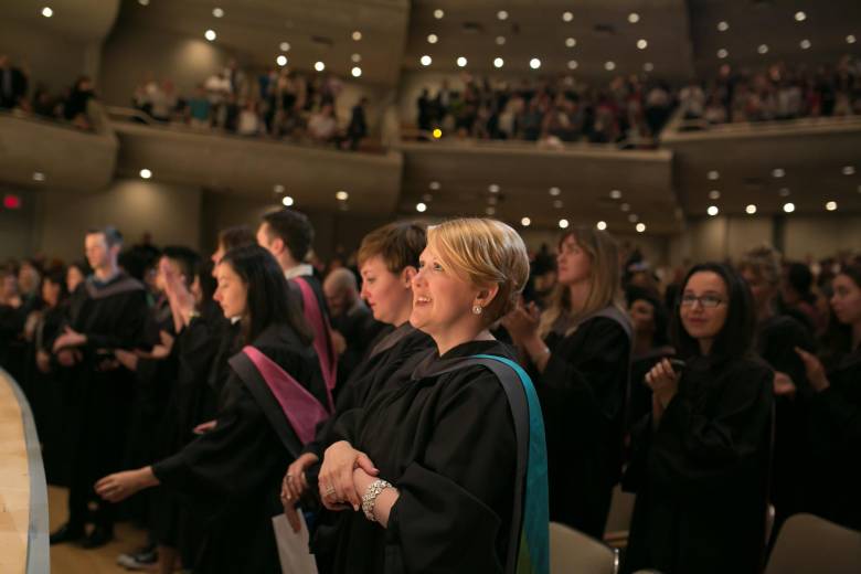 Photo from past Convocation ceremony shows an audience of graduands standing, smiling, clapping, wearing their robes. The photo focuses on one individual smiling towards the stage.