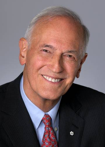 Photo of an older white man with grey-white hair with brown eyes smiling and wearing a navy blazer and light blue shirt and red and blue tie.