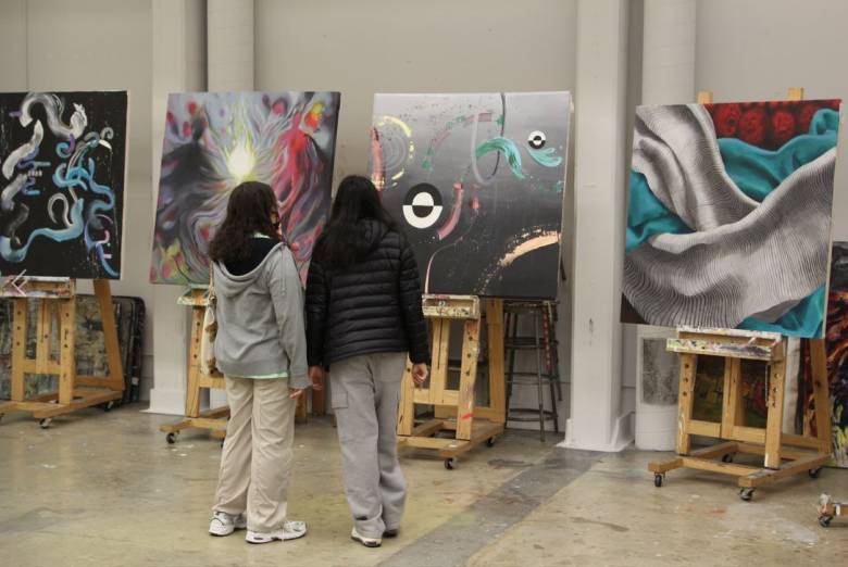 Two women looking at several colourful canvases on easels (back view).
