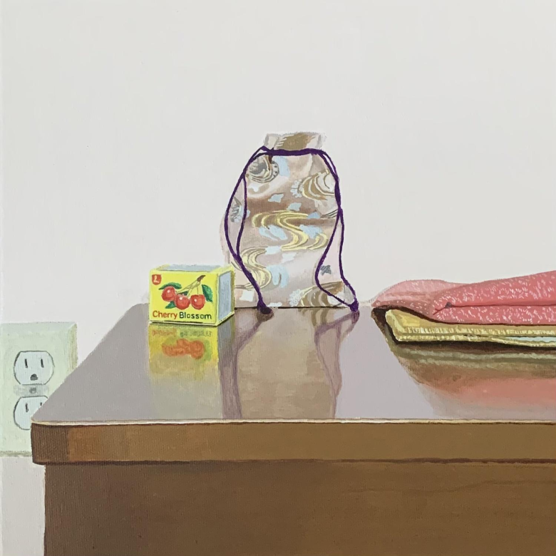 A still life painting of a brown desk with objects on it including a violet coloured silk bag, two folded textiles and a small yellow box with three cherries on it with the words “Cherry Blossom”.