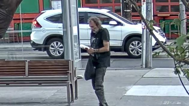 A security camera image of a white man with facial hair wearing a black t-shirt and faded black jeans.