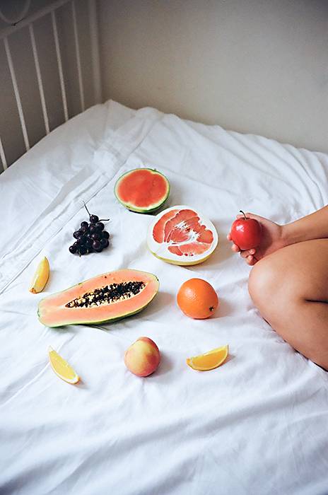 Photographic image of an assortment of fruit and naked leg on a bed with white sheets