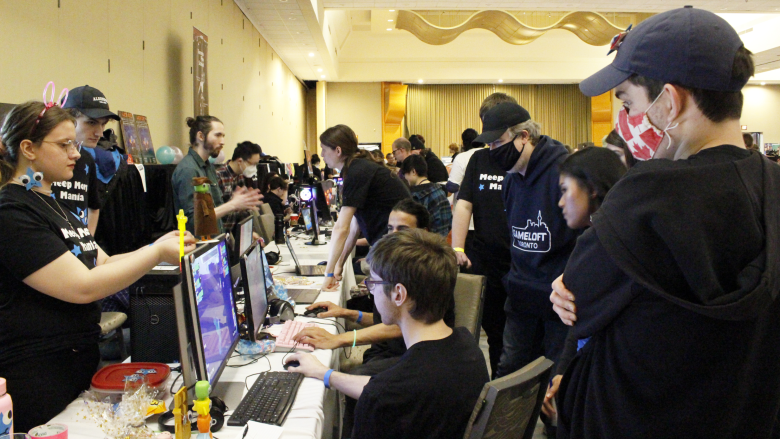 Image from Level Up Showcase, courtesy of Dr. Emma Westecott. Many people on either side of a table full of computers with games on them.