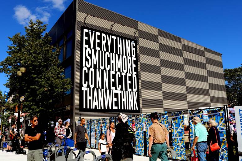 Image graphic showing public art installation by Hiba Agdullah, a large billboard style sign on the outside of a building says "Everything is much more connected than we think"