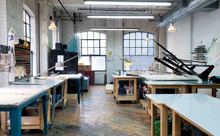 Printmaking studio with wooden floors, wooden desks, printmaking equipment and large grated windows. 