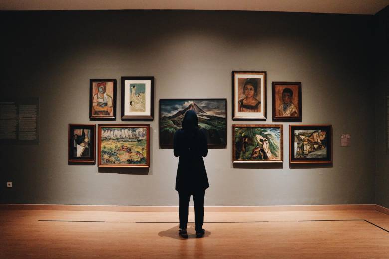 A person stands in a gallery in front of paintings on a wall.