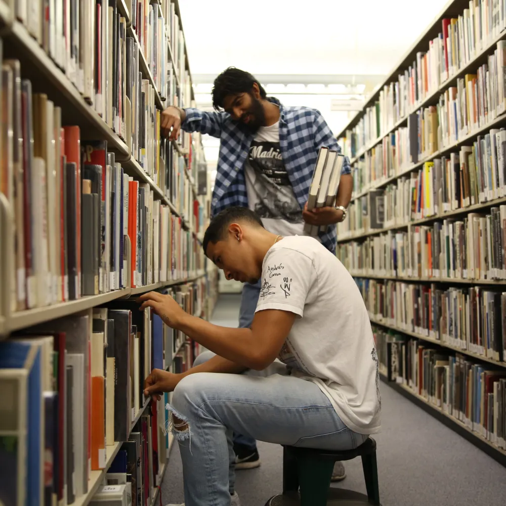 One person sitting and another standing inside library stacks.